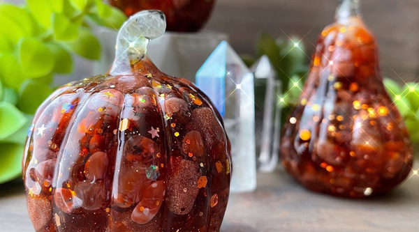 two orange resin pumpkins with glitter and encapsulated stones sitting on a wooden surface amongst crystals and greenery