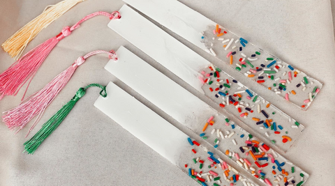 four resin bookmarks with sprinkles in one end and solid white on the other laying on top of a stone background all with tassels of different colors