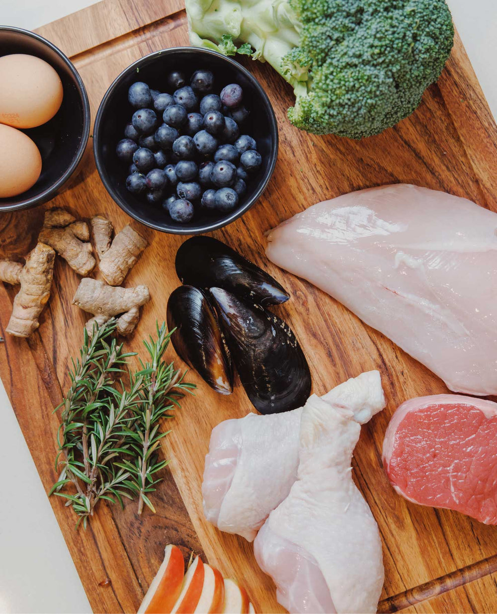 A variety of fresh ingredients on a wooden board, including eggs, blueberries, broccoli, and meats.