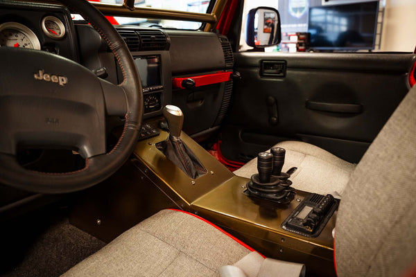 A close-up of the interior of a red Jeep Wrangler, showing the steering wheel, dashboard console, and front seats. The Jeep has aftermarket seats with harnesses, a custom steering wheel, a large touchscreen in the center console, and various switches and gauges.
