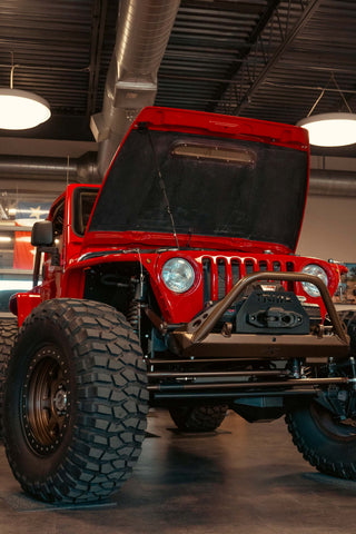 A red Jeep LJ with a flame red and highland bronze paint job with its hood open in a garage. The Jeep has aftermarket bumpers, a winch, lifted suspension, and large tires. It also has a custom cage and interior with PRP seats. Tribe Offroad logos are visible throughout the vehicle.