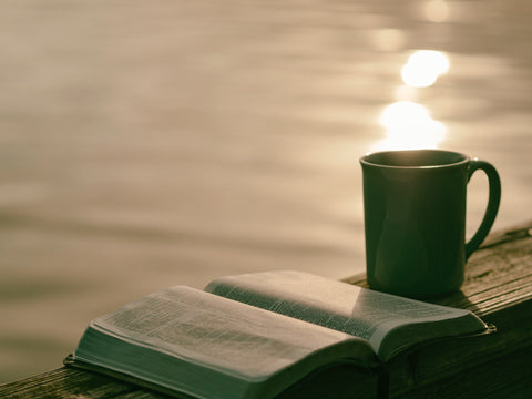 An open book and a mug on a wooden surface looking over the view of a lake.