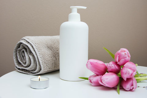 A white cosmetic pump bottle placed on a white surface alongside a small lit candle, a rolled-up brown bath towel, and a bouquet of pink tulips.