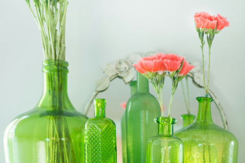 An arrangement of various bottles made out of green-tinted glass modified into vases used to hold pink flowers.