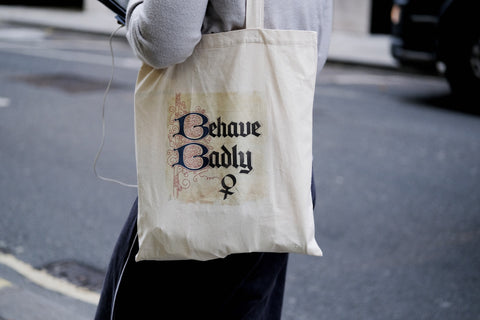 A person carrying a cotton tote bag decorated with the words “Behave Badly” in a black Gothic font on their shoulder.