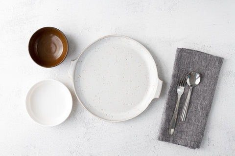 A reusable plate and two reusable bowls arranged alongside a stainless steel fork and spoon on a grey cloth napkin.