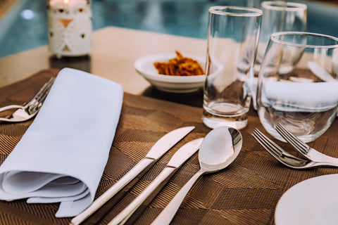 A table set with stainless steel cutlery and drinking glasses on top of a brown placemat.