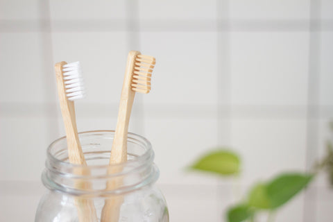 Two bamboo toothbrushes inside a glass jar, placed in front of a white-tiled bathroom wall.