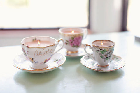 Three vintage porcelain teacups with flower patterns and matching plates that have been upcycled into candles with pink wax.