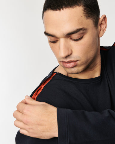Ein Mann mit dunkelbauen Sweatshirt, er hat die Hand auf der Schulter und zeigt die Druckknofleiste