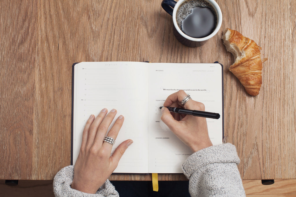 Mesa vista desde arriba con unas manos escribiendo en un diario junto a un café y un croissant.