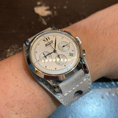 A man's wrist is shown wearing a Tissot wristwatch with a cream white face, silver hands, and a silver bezel. The watch is paired with a grey leather bund style watch strap, which has a textured surface and a silver metal buckle. 