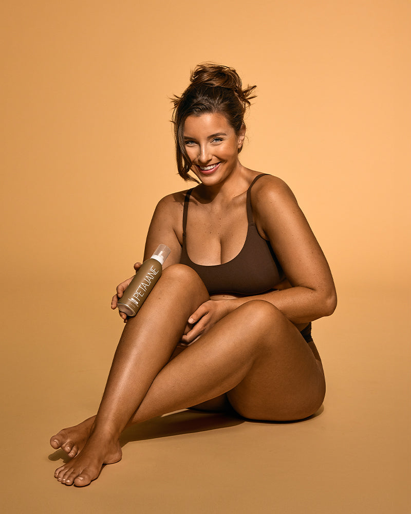 Woman in brown swimsuit holding a skincare product against a warm-colored background.