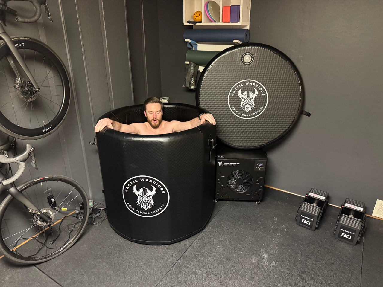 Man in a cold plunge tub beside exercise equipment in a home gym setting.
