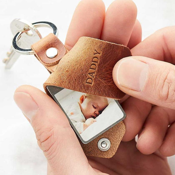 A brown leather keyring with a photo of a baby inside and the word 'Daddy' in debossed lettering, held in a man's hand