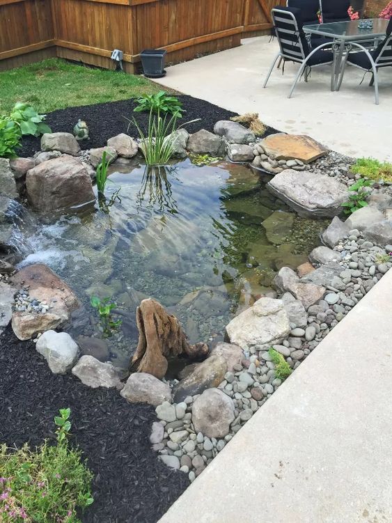 Small backyard pond set in the corner of a concrete patio filled with rocks and crystal clear water