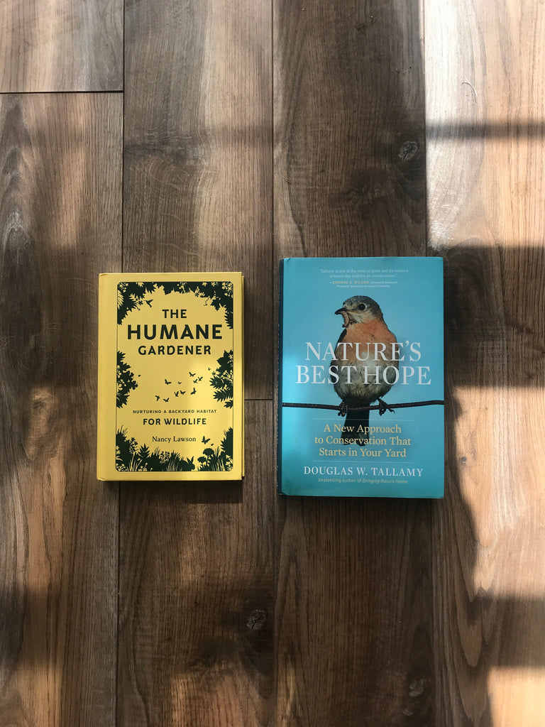 A yellow book (The Humane Gardener) and blue book with a bird (Nature's Best Hope) lie on a wooden floor. A window is casting light and shadow all across the books and floor. 