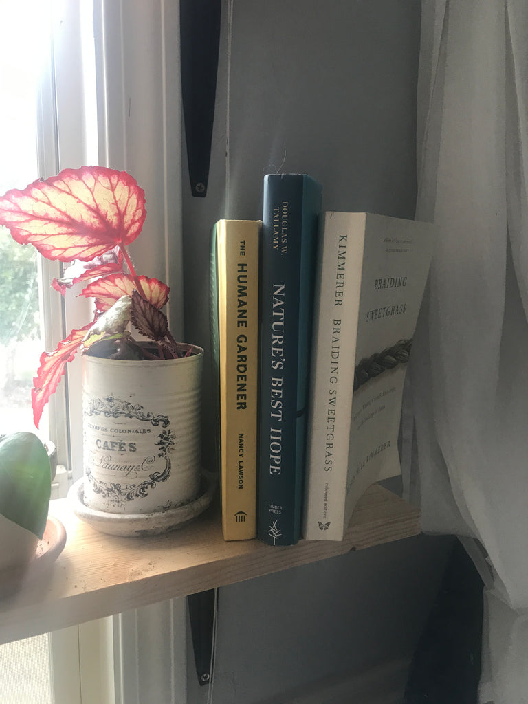 Three books (The Humane Gardener, Nature's Best Hope, and Braiding Sweetgrass) sit on a light wooden shelf next to a reddish plant in a white tin can