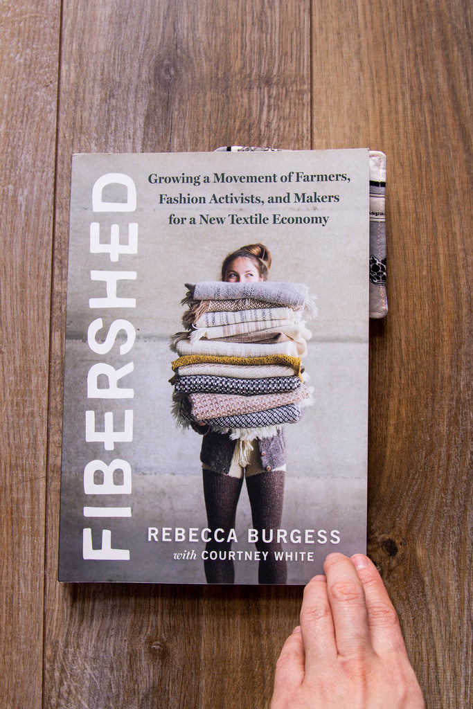 A straight on image of a book laying against a dark wood floor. The title of the book is along the left side and reads "Fibershed" in white letters against a grey background. On the center of the cover is a white woman holding a variety of hand knit and woven fabrics with her head peeking up. Above her head reads " Growing a movement of farmers, fashion activists, and makers for a new textile economy." At the bottom of the cover it reads "Rebecca Burgess with Courtney White" in white lettering