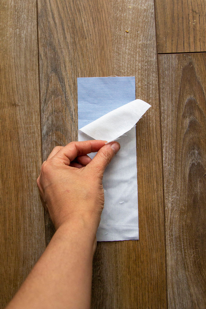 A blue fabric rectangle sits on top of a dark wooden floor with the muslin rectangle being placed over the blue fabric by a white hand. 