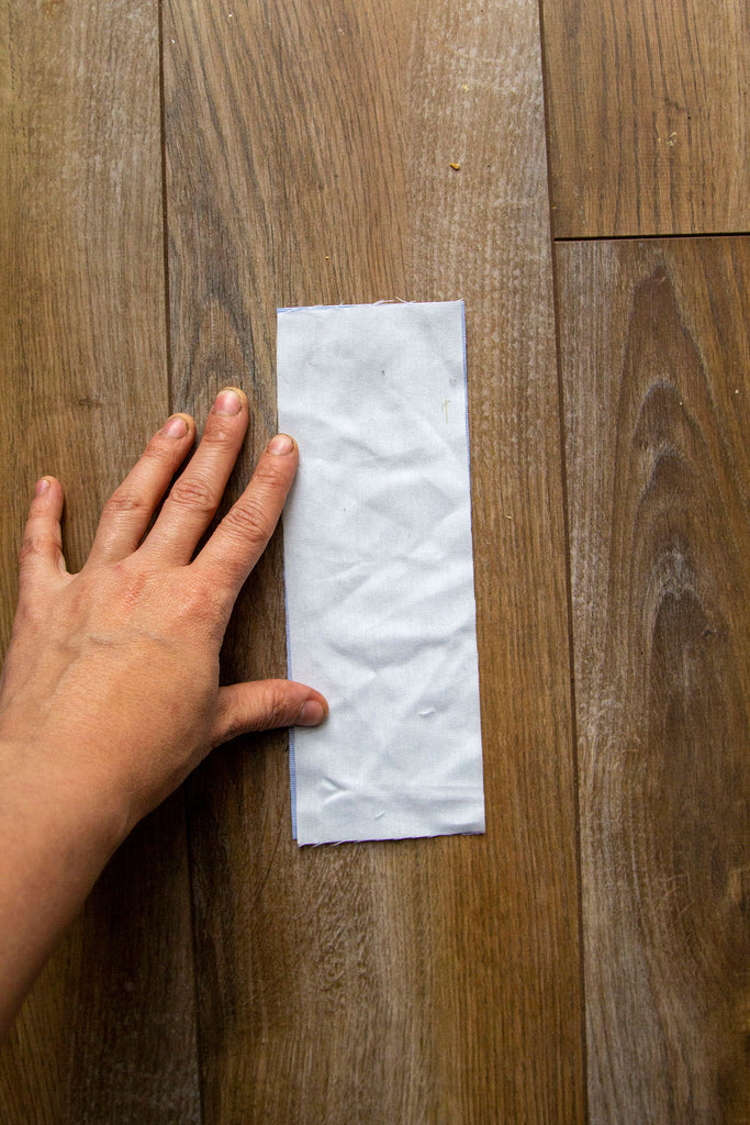 A blue fabric rectangle sits on top of a dark wooden floor with the muslin rectangle completely covering the blue fabric with a white hand holding everything in place to the left. 