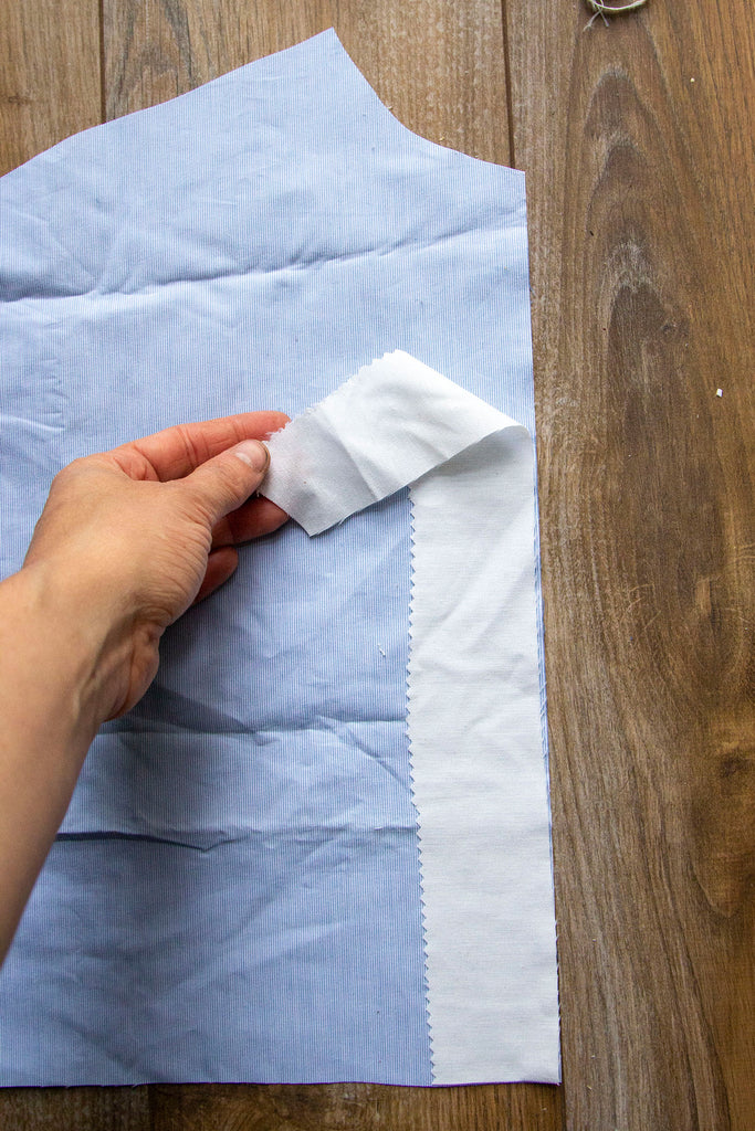 A pair of white hands is placing the rectangular muslin piece on top of the blue fabric along a straight edge on the right hand side. The finished edge of the interfacing is facing towards the inside of the blue fabric.  