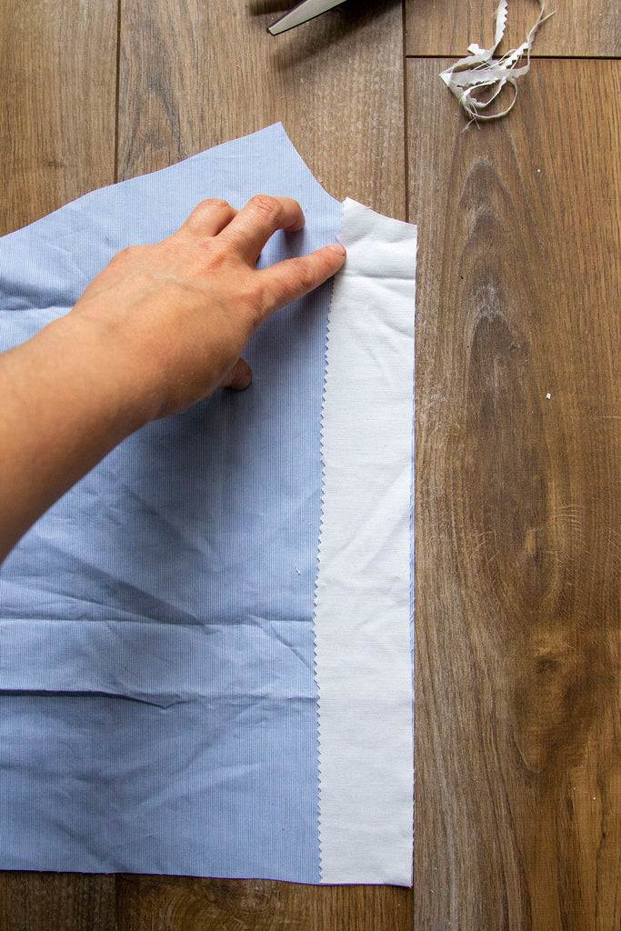 A pair of white hands has placed the rectangular muslin piece on top of the blue fabric along a straight edge on the right hand side. The finished edge of the interfacing is facing towards the inside of the blue fabric.  