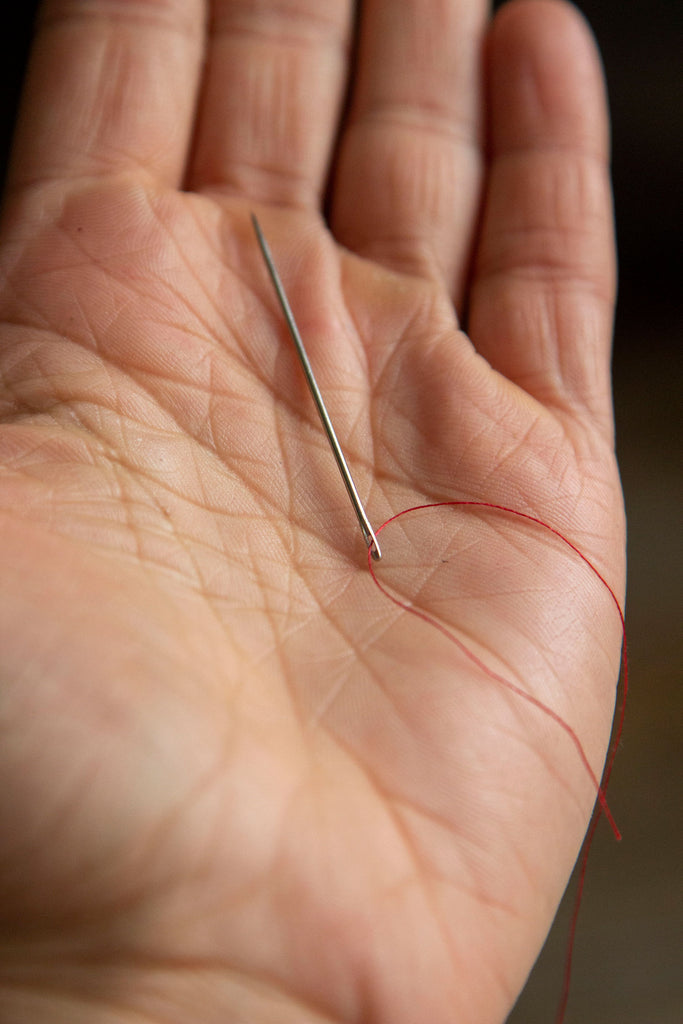 A metal needle sitting on a white palm with a single red thread running through its eye. 