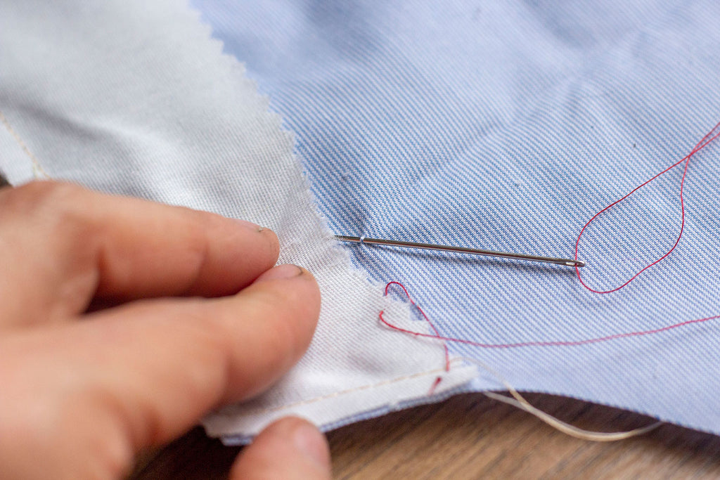 The single red thread is attached to the metal needle which is picking up one thread on the blue garment. A white hand is pulling the muslin interfacing to one side to get a better view of the needle picking up a singular thread. 