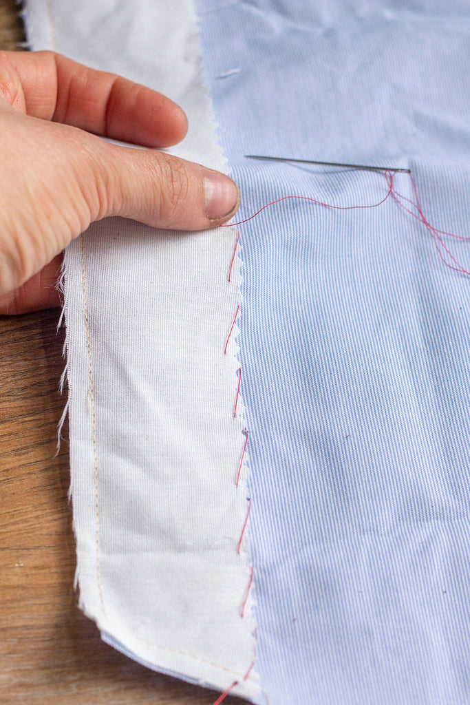 An in progress shot of the red thread creating a blind stitch on the wrong side of the garment. A white hand and thumb are being used to evenly space the stitches along the muslin interfacing. 