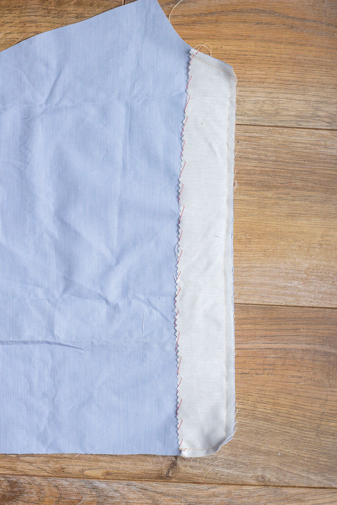 A straight on shot of the wrong side of the blue garment with muslin interfacing along the right side of the fabric. There are a line of red stitches seen against the muslin interfacing. Part of the blue garment is cropped from view.