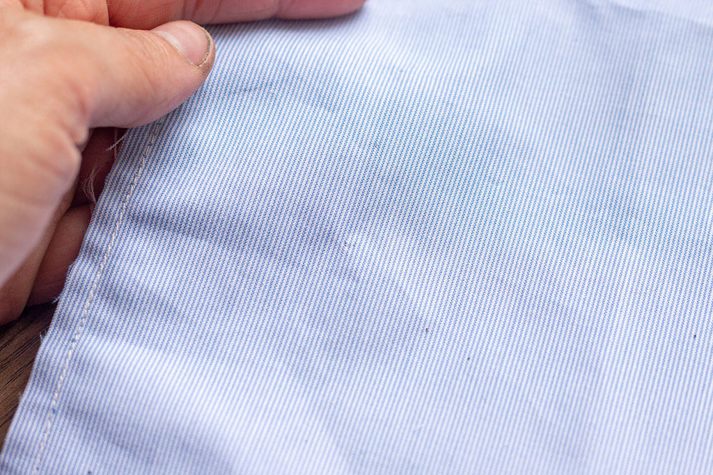 A close up of the blue garment being held in a white hand where the red threads are not visible except for maybe a small pin prick texture on the fabrics surface. 