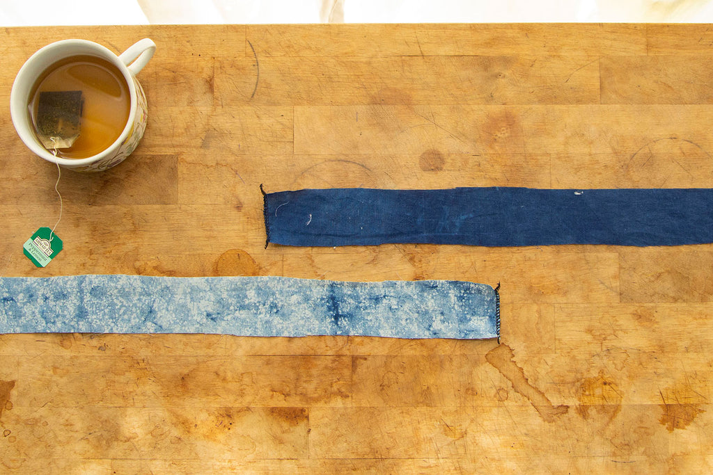 Two different blue colored waist ties are lying on a wooden cutting board, each with a short end serged. A cup of tea sits in the upper left hand corner. 