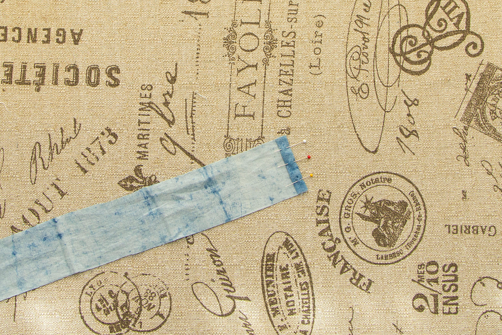 A close up of one end of a blue casing with the short end folded back twice and pinned into place. It sits on a light brown ironing board covered in dark brown french writing