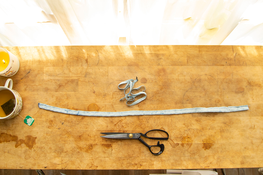 A light blue waist tie folded in half lengthwise with half of its seam trimmed and a pair of black scissors sitting under it. IT sits on a wooden chopping block with a candle and cup of tea in the upper left hand corner. 