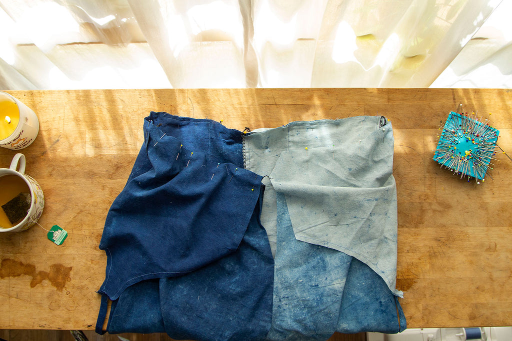 The back opening of the jumpsuit is seen with the bodice and pants having been attached with pins. Dappled light hits the wooden cutting board