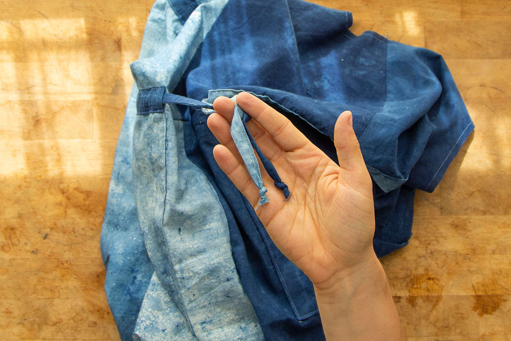 A white hand showcases both ends of the waist ties tied into small knots. The blue jumpsuit is seen in the background. 