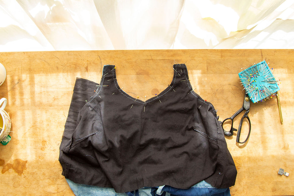 A flat lay of a black bodice lining pinned along the front neckline, strap opening and underarm of the jumpsuit bodice. A pair of scissors and pins is in the right hand corner