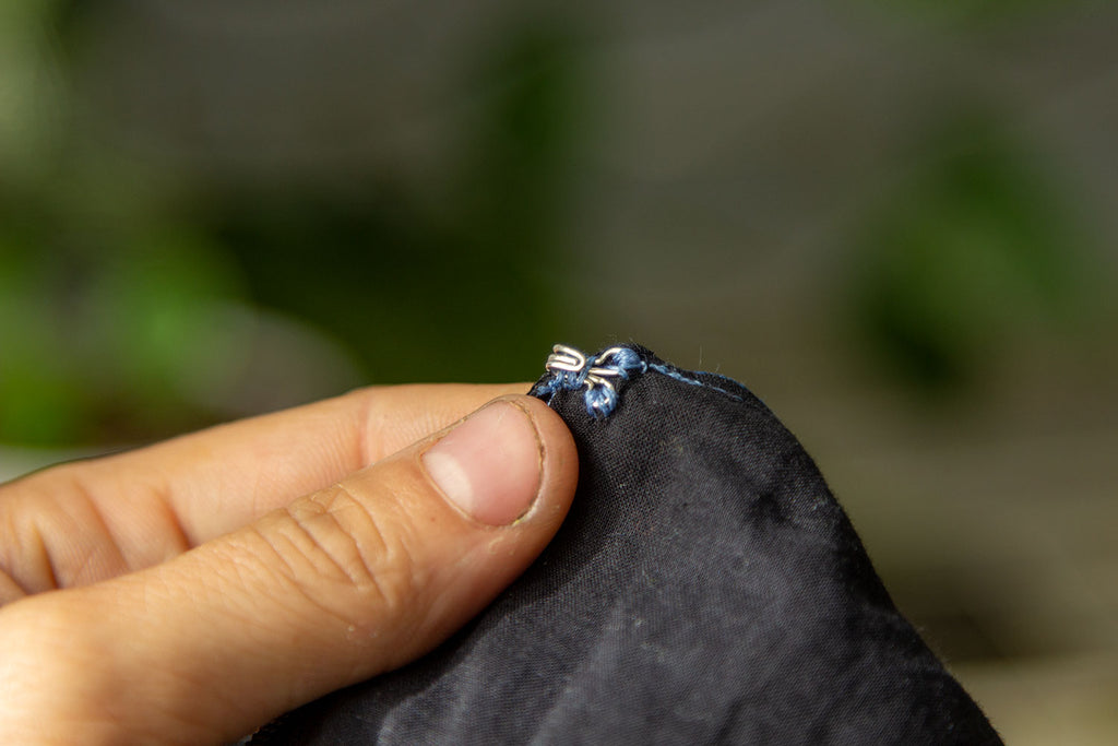 A white hand holds up a small silver hook that has been sewn with blue thread to one side of the inner jumpsuits black lining. 