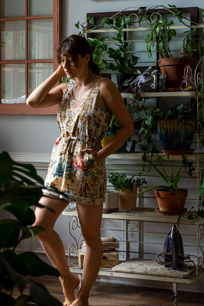 A white woman is standing in front of a white bakers rack filled with many plants. She is wearing a beige colored romper with thick straps and a floral print
