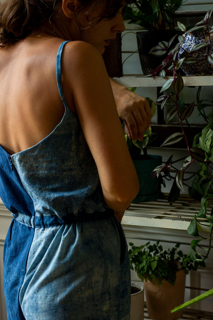 A white woman's back and part of her face is shown as she rests her arms on a white bakers rack filled with plants. She is wearing a dual blue colored jumpsuit with an invisible zipper down the back