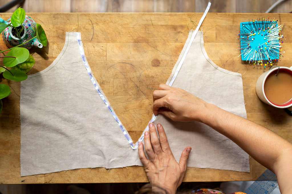 White satin ribbon is being pinned in place by a pair of white hands to a cream colored linen back bodice neckline