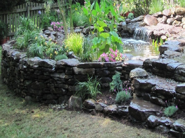 A pond built on top of a flat rock stacked retaining wall with a soft waterfall feature to the side. 