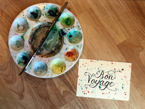 A paint palette filled with watercolor paints, a paint brush and a card splattered in colorful paint droplets that reads "Bon Voyage"