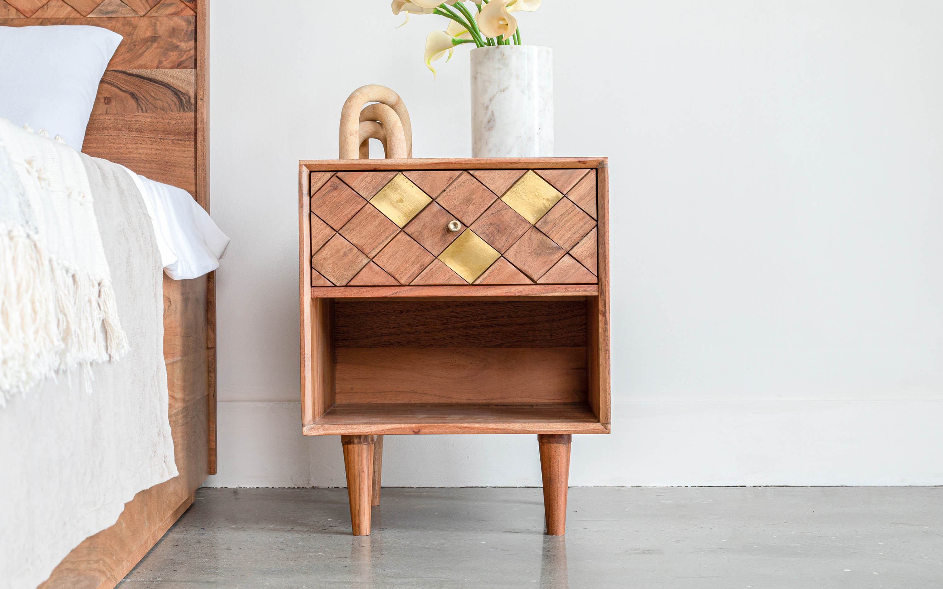 The bedside's ambiance is elevated with an artisan side table, its wooden craftsmanship adorned with brass geometric inlays, complemented by an ornamental vase that adds a flourish of botanical elegance