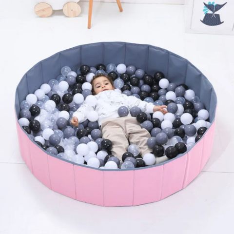 Enfant allongé détendu dans une piscine à balles hexagonale rose et grise, entouré de balles noires, grises et blanches, avec un décor de chambre d'enfant en arrière-plan.
