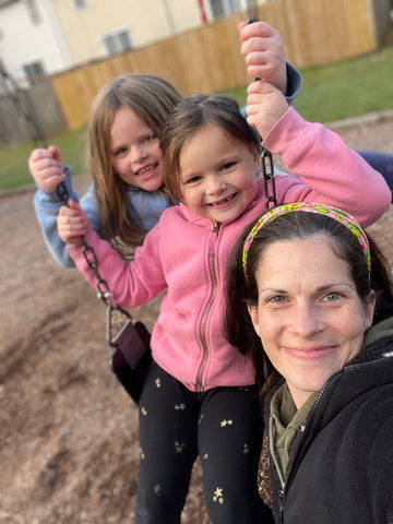 Mom and 2 daughters at a playground. Girl in pink sweatshirt seated on a swing with mom in front of her to the right and sister behind her to the left wearing a light blue sweatshirt.