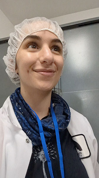 Person smiling, wearing a hairnet, white coat, and blue lanyard in a medical or laboratory setting.