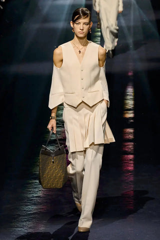 Model in cream cold shoulders suit vest with pleated skirt worn over loose-fitted trousers