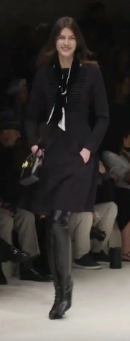 Model in runway in a long-sleeved, black midi dress with thigh-high boots and a black and white scarf